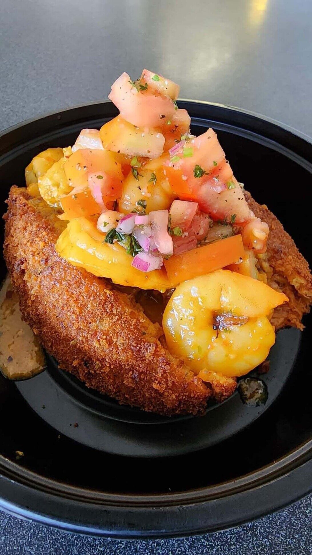 Alcapurria topped with shrimp, melted cheese, and fresh pico de gallo in a black bowl